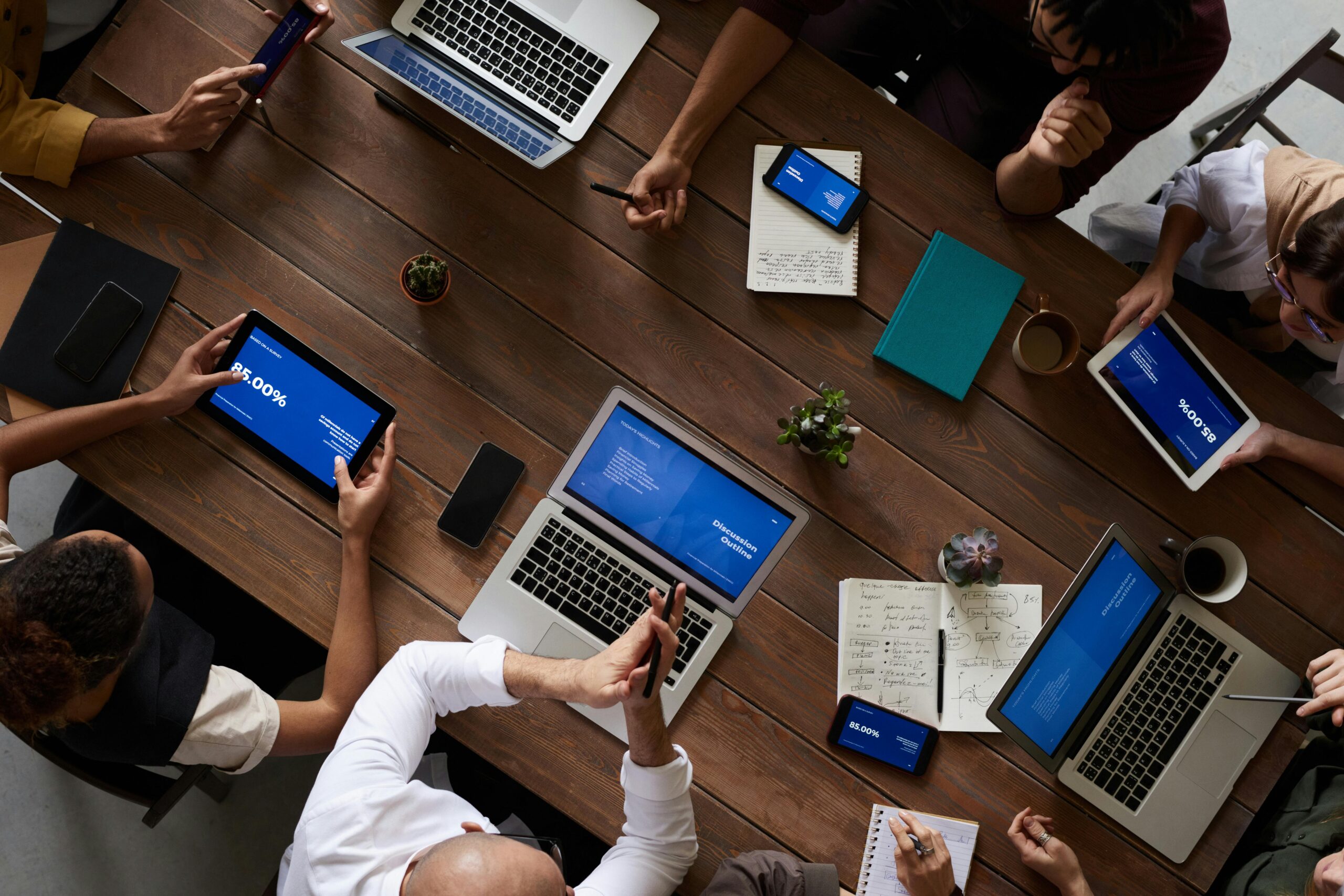 Een vergadertafel van bovenaf. Er liggen meerdere notitieboekjes opengeslagen, en er staan meerder laptops open. Ook staan er kopjes koffie en een paar plantjes